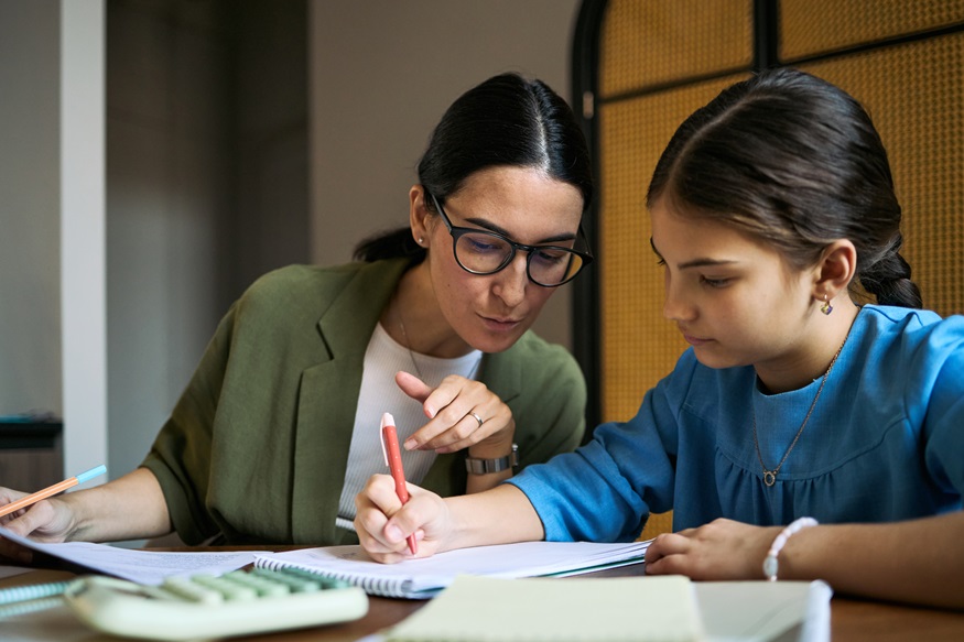 Middle aged woman guiding schoolgirl while writing in workbook during tutoring session. Education support concept for homeschooling, private lessons, parenting campaigns, medium shot