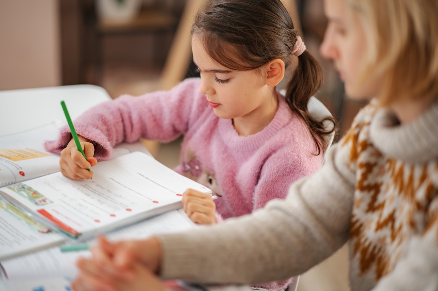 Girl diligently writing on notebook with pencil, mother supporting during homework